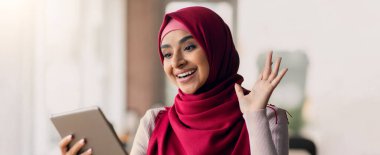 Kırmızı tesettürlü genç bir kadın tabletine video çağrısı yaparken gülümsüyor. Aydınlatılmış parlak bir mekanda otururken mutlu ve meşgul görünüyor..
