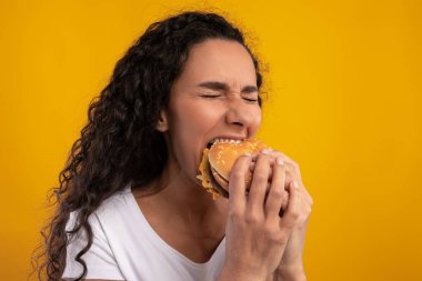 Neşeli bayan parlak sarı bir arka planda duruyor, büyük bir burgeri ısırmaya hazır..