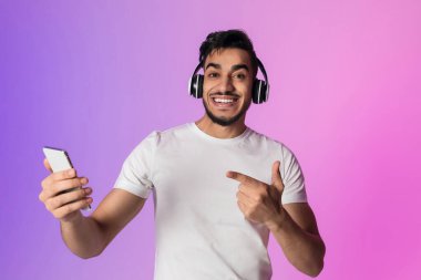 Garip genç Arap adam kablosuz kulaklık takarken geniş bir gülümseme takıyor. Renkli neon ışıklı bir ortamda müzik dinleyerek akıllı telefonunu işaret ediyor. Mobil uygulamaları desteklemek için mükemmel.