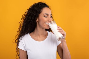 Mutlu genç bir kadın sarı-turuncu bir duvarla stüdyoda duruyor, bardaktan süt içiyor ve gülümsüyor. Sağlıklı, taze organik süt ürünlerini seviyor, neşe ve canlılık saçıyor..
