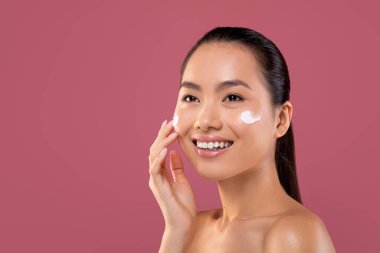 Güzel bir Çinli kadın yanaklarına nemlendirici sürerken gülümser. Mor bir stüdyoda çalışıyor ve yeni bir güzellik ürünü denemek için sabırsızlanıyor..