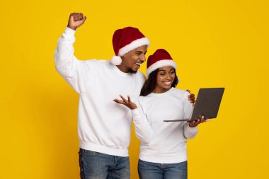 Neşeli bir çift Noel Baba şapkaları ve beyaz kazaklar giyiyor. Bir dizüstü bilgisayarla etkileşime geçiyorlar, tatil sezonunda birlikte neşeli bir anı paylaşıyorlar, sarı bir arka planda..