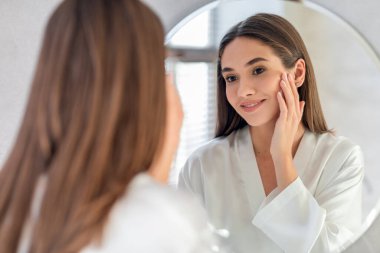 Çekici genç bir kadın parlak bir banyoda hazırlanırken aynadaki yansımasına gülümsüyor. Doğal güzelliğinin tadını çıkarıyor ve cilt bakımına odaklanıyor. Kendine güveni tam..