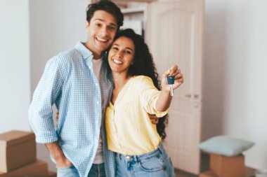 İki gülümseyen birey, taşınan kutularla dolu parlak bir salonda yan yana duruyorlar. Gururla ellerinde anahtarlarla yeni evlerini kutluyorlar..