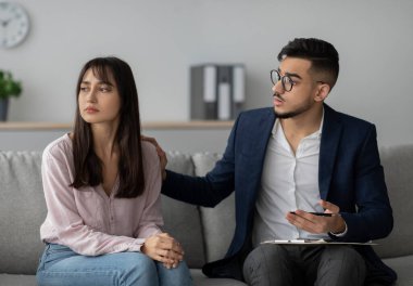 Orta doğulu üzgün bir bayan, modern bir klinik ortamında psikoterapist onu rahatlatırken bir koltukta oturur. Atmosfer profesyonel bir danışmanlık ortamını yansıtıyor..