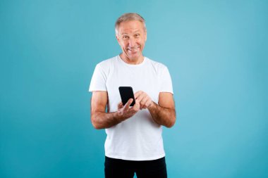 Olgun bir adam mavi arka planlı bir stüdyoda duruyor, akıllı telefonunu kullanırken gülümsüyor. Meşgul ve neşeli görünüyor, muhtemelen sosyal medyada geziniyor ya da internette sohbet ediyor..