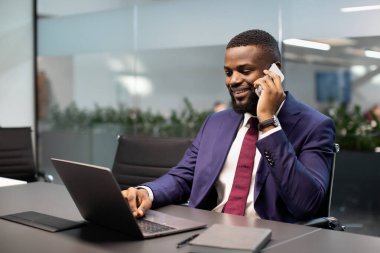 Başarılı Afro-Amerikan genç iş adamı, pahalı mavi takım elbiseli, ofiste dizüstü bilgisayarla çalışıyor, kişisel asistanı ya da iş ortağıyla telefon görüşmesi yapıyor ve gülümsüyor.