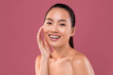 Güzel bir Asyalı bayan mor bir stüdyo geçmişine karşı üstsüz poz veriyor. Gülümsüyor ve yüzüne dokunuyor. Sağlıklı, ipeksi tenini gösteriyor ve anın tadını çıkarıyor..