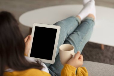 Bin yıllık Arap bir kadın boş ekranlı dijital tablet kullanarak evdeki kanepesinde rahatça oturuyor. Elinde bir fincan çay tutuyor, boş zamanlarının tadını çıkarıyor..