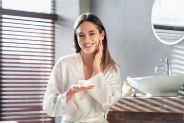 Genç bir kadın yüzüne nemlendirici krem sürerken kameraya gülümsüyor. Parlak bir banyoda, bornoz giyiyor, gündüzleri güzellik rutininin tadını çıkarıyor..