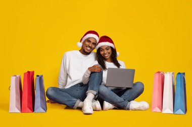 İki genç yetişkin, renkli alışveriş torbalarıyla yerde oturuyor. İnternetten alışveriş yaparken Noel Baba şapkası takıyorlar. Noel ruhunu ve neşesini somutlaştırıyorlar..