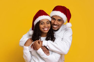 Mutlu bir çift birbirlerini kucaklarken kırmızı Noel şapkaları giyer. Parlak gülümsemeleri ve şenlikli kıyafetleri neşeli bir tatil atmosferi yaratıyor. Arka plan canlı bir sarı..