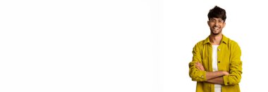 Genç bir adam kollarını kavuşturup gülümseyerek ayakta duruyor. Şık gözlükleri var ve beyaz bir gömleğin üzerine sarı bir ceket giyiyor. Arka plan sade, neşeli varlığını arttırıyor..