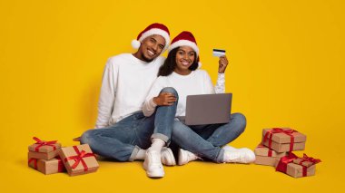 Neşeli bir çift internette alışveriş yaparken yerde Noel şapkası takıyor. Etrafları hediye paketleriyle çevrili. Bayram sezonu için heyecanlarını sergiliyorlar..