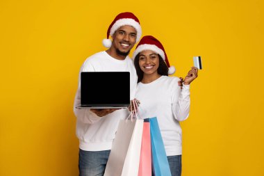 Neşeli bir çift dizüstü bilgisayar ve alışveriş torbaları tutarken Noel Baba şapkası takıyor. Gülümsüyorlar ve Noel alışverişinin neşeli ruhunu yansıtan bir kredi kartı gösteriyorlar..