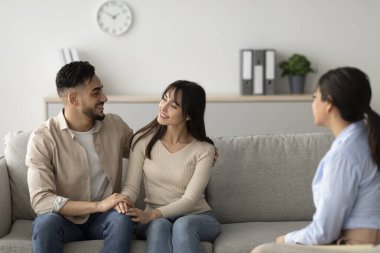 Neşeli bir Arap çift danışman ofisindeki bir kanepede oturup birbirlerine bakarken birbirlerine gülümsüyorlar. Profesyonel bir psikolog oturumlarına rehberlik eder, açık iletişimi teşvik eder..