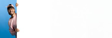 Pembe kazak ve şapka giyen neşeli bir adam iki eliyle boş bir tabela tutuyor. Canlı bir mavi arkaplana karşı duruyor, bir mesajı ya da duyuruyu paylaşmaya hazır..