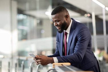 Takım elbiseli Afrikalı Amerikalı CEO akıllı telefonlarda iş telefonu uygulaması kullanıyor. Borsa ve hisse senedi ticareti yapan siyahi işadamı ofiste mola veriyor. Kopya alanı olan panorama.