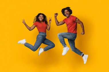 İki genç yetişkin sevinçle havada zıplıyor, heyecan ve enerji gösteriyorlar. Eşleşen kırmızı gömlekler ve açık renk kot pantolon giyiyorlar, barış işaretleri ve neşeli ifadeler sergiliyorlar..