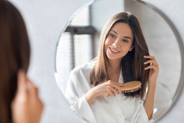 Çekici genç bir kadın güzel saçlarını aynanın önünde zevkle tarar. Yumuşak beyaz bir bornoz giyer ve parlak bir banyo ortamında yansımasına gülümser..