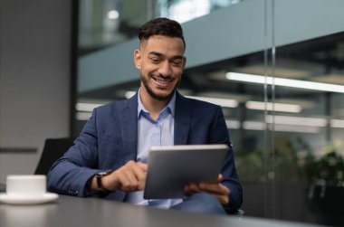 Neşeli bir Orta Doğulu CEO, modern bir ofiste masada oturuyor ve tabletindeki son iş telefonu uygulamasını kontrol ederken gülümsüyor. Güzel bir takım elbise giymiş, anın tadını çıkarıyor..