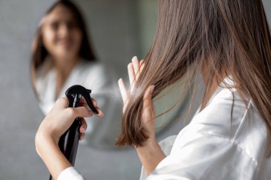 Genç bir dişi banyoda duruyor, aynadaki yansımasına bakarken saç spreyi sıkıyor. Yeni saç stili ürününü test ediyor ve saç modeline odaklanıyor..