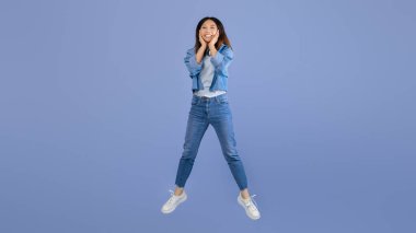 Gündelik giyinmiş genç bir kadın mavi bir zemine sevinçle zıplıyor. Geniş bir gülümseme sergiliyor, bu canlı anda heyecanını ve mutluluğunu gösteriyor..