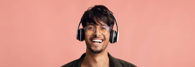 Genç bir adam kulaklık takarak eğleniyor. Yüzünde büyük bir gülümseme var ve yumuşak pembe bir arka planın önünde neşeli ve canlı bir atmosfer yaratıyor..