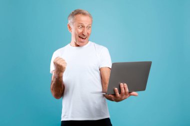 Neşeli son sınıf öğrencisi mavi bir stüdyoda dizüstü bilgisayarını kullanırken heyecan gösteriyor. Kişisel ya da profesyonel bir başarıyı neşeli jestler ve büyük bir gülümsemeyle kutlar..