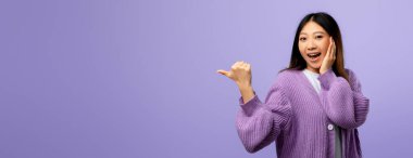 Uzun saçlı genç bir kadın mor bir kazak giyerken gülümsüyor. Heyecanlı bir şekilde sağını işaret ediyor. Gün ışığında tarafsız mor bir arkaplan sergiliyor..