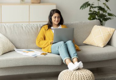 Heyecanlı bir Arap kadın koltukta rahatça oturuyor ve dizüstü bilgisayarı üzerinde çalışıyor. O bir kulaklık takıyor ve aktif olarak yazıyor. Evraklar etrafa dağılmış, online işlerle ilgilendiğini gösteriyor..