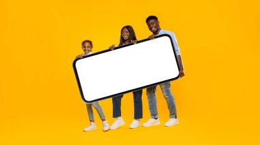 Üç kişilik aile, parlak sarı-turuncu bir arkaplan karşısında boş ekranlı büyük ve ağır bir akıllı telefon tutuyor. Bu sahne mobil uygulama tanıtımı ya da yaratıcı reklamcılık için mükemmel..