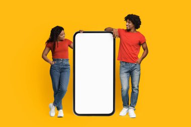 Genç bir çift, canlı sarı bir zemin üzerinde büyük bir boş tabela sunarak yan yana duruyor. Neşeli ve rahat görünüyorlar. Kırmızı gömlekler ve kot pantolon giyiyorlar..