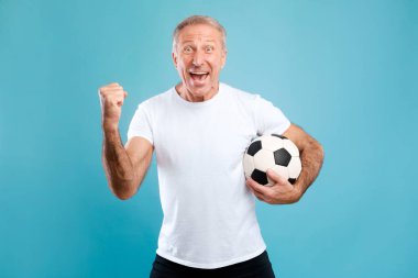 Olgun adam enerjik bir şekilde poz veriyor, elinde bir futbol topu tutuyor ve kazanan bir jest yapıyor. Mavi bir stüdyonun önünde dikilerek bir gol attığı için çok heyecanlı..