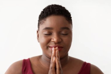 Kısa saçlı, çekici, şişman siyahi bir kadının meditasyon yaparken çekilmiş portresi. Gözleri kapalı ve elleri namaste jesti yapıyor. Huzurlu atmosfer stresi azaltıyor..