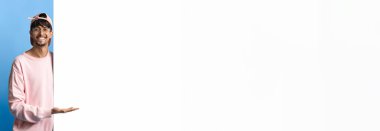 Pembe kazak ve kep giyen neşeli bir adam mavi arka planda duruyor. Gülümsüyor ve sanki izleyicileri dikkat çekmeye davet ediyormuş gibi hareket ediyor..