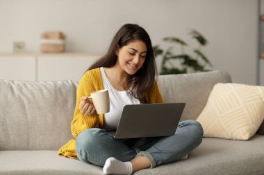 A happy arab female freelancer sits on her sofa, sipping coffee from a cup. She is engaged with her laptop, working online in a bright and inviting living room space.