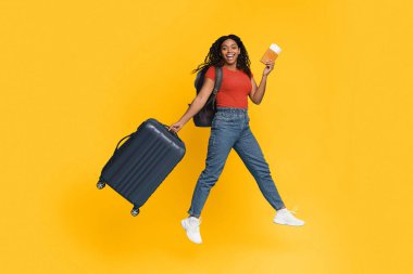 Gülümseyen genç bir kadın elinde bir bavul ve içecek tutarken sevinçle zıplıyor. Günlük kıyafetler ve sırt çantası giyiyor. Parlak sarı arka plan sahneye enerji katıyor..