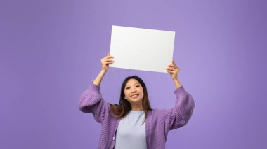 Bir kadın havada boş bir tabela tutarken parlak bir şekilde gülümsüyor. O, rahat bir lavantalı kazak giyiyor. Parlak mor arka plan, sahneye neşe katıyor..