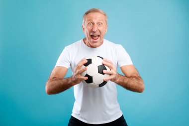 Elinde futbol topu olan neşeli, olgun bir adam mavi bir arka plana karşı duruyor. En sevdiği takımı desteklemek ve oynamak için heyecanlı. Onun coşkusu spor ve eğlence faaliyetlerinin neşesini vurguluyor.