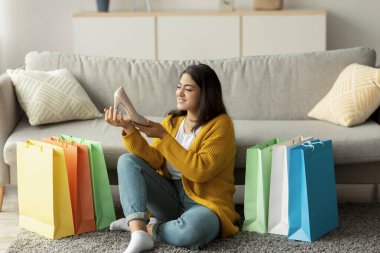 Heyecanlı bir Arap kadın elinde yeni bir ayakkabıyla evde yerde oturuyor. Başarılı uzaktan alışveriş deneyimini yansıtırken etrafını renkli alışveriş torbaları sarmış..
