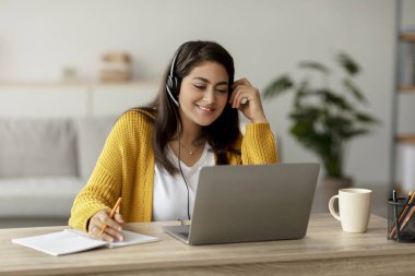 Ev ortamında dizüstü bilgisayarını kullanırken genç bir Arap kadına parlak bir gülümseme. Kablosuz bir kulaklık takıyor ve yanında bir defter var..