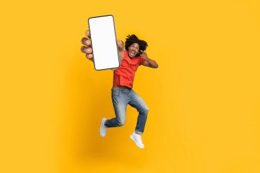 Kırmızı gömlek ve kot pantolon giyen neşeli bir adam elinde boş bir akıllı telefon ekranı tutarak havaya zıplıyor. Parlak sarı arka plan, yakalanan neşeli havayı arttırır..