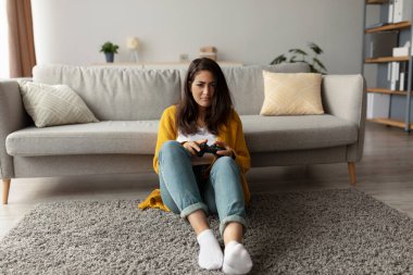 Ciddi bir Arap kadın oturma odasında halıya oturmuş, elinde joystick ile video oyunları oynamaya odaklanmış. Mekan temiz ve davetkar, arka planda hafif ışıklandırma ve kanepe var..