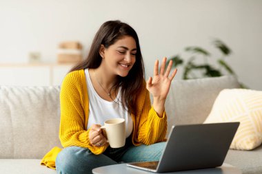 Genç bir Arap bayan, elinde kahve fincanı, kameraya el sallarken gülümseyen bir şekilde kanepesinin konforundan laptopundan arıyor. Oturma odası aydınlık ve davetkar..