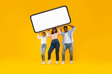 Üç kişilik siyah bir aile ağır, büyük bir akıllı telefon ve boş bir ekran tutuyor. Parlak sarı turuncu bir stüdyoda gülümserler, mobil bir uygulama veya web sitesi fırsatını tanıtırlar..