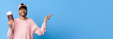 Genç bir adam mavi bir arkaplanda duruyor, tek eliyle gülümsüyor ve işaret ediyor. Öte yandan elinde bir belge tutuyor ve pembe bir kazak giyiyor. Aynı kepten takıyor. Heyecanı gösteriyor..