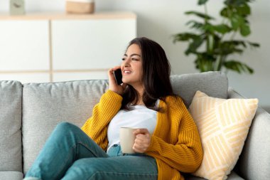 Arap bir kadın oturma odasındaki koltukta dinleniyor, kahvesini yudumluyor ve cep telefonuyla konuşuyor. Arkadaşça sohbet ediyor, evde boş vaktinin tadını çıkarıyor..