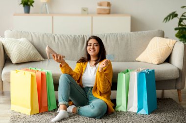 Sıcak bir ev ortamında, neşeli bir Arap kadın renkli alışveriş torbalarıyla çevrili yerde oturuyor. Elinde bir kredi kartı ve yeni bir ayakkabı var. İnternetten alışveriş yaparken gülümsüyor..