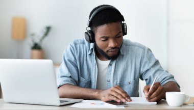 Kulaklıklı konsantre olmuş Afro-Amerikalı adam video konferansı yoluyla yabancı dil eğitimi alıyor ve not defterine, panoramada kopya alanı olan notlar alıyor.